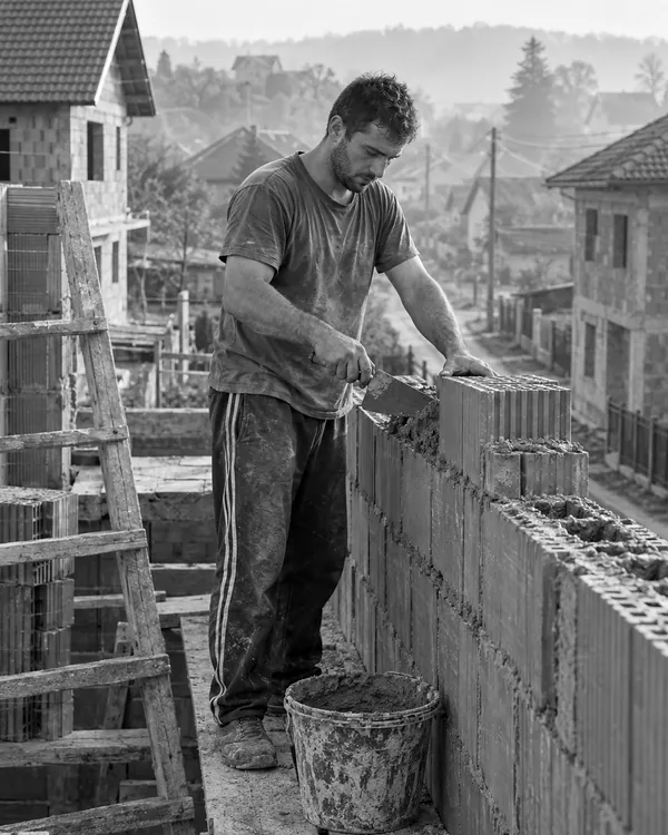 A young worker in an old tracksuit, with no helmet or safety gear, lays blocks on an unfinished house in a village.
