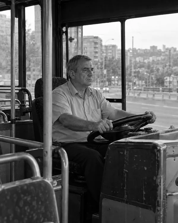 A city bus driver in a light blue shirt drives in the early morning, both hands on the wheel, looking ahead.